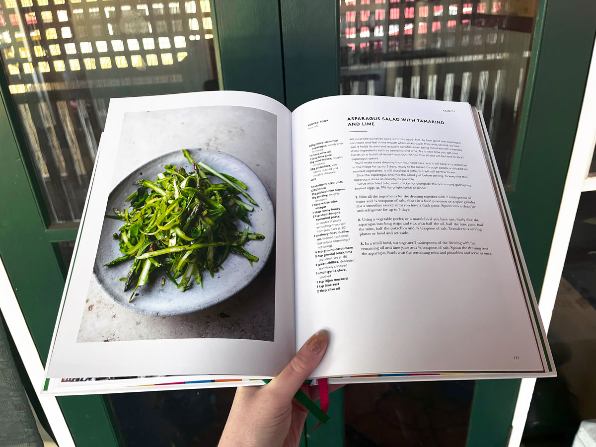 A book being held open at a page showing an image and recipe for asparagus salad with tamarind and lime