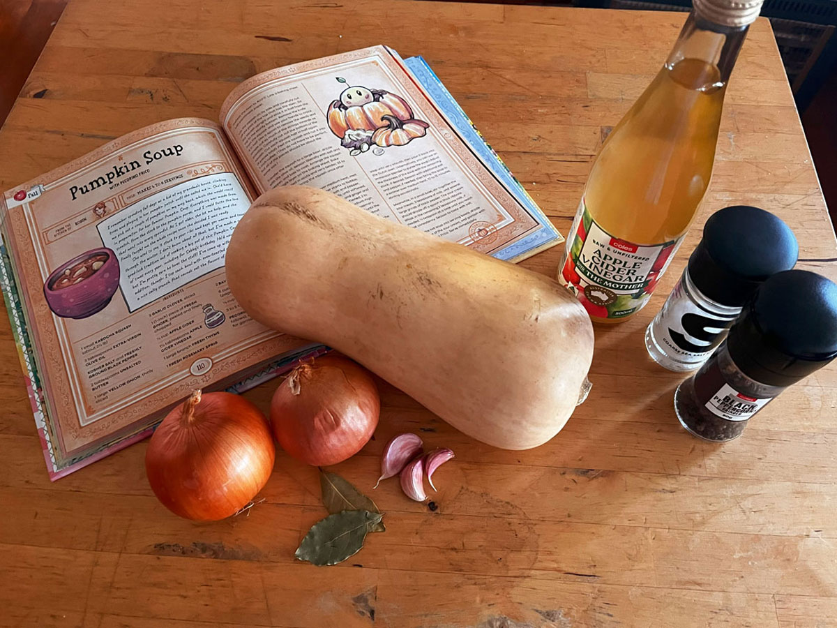 Ingredients (a butternup squash, two onions, some bay leaves, cloves of garlic, a bottle of apple cider vinegar, salt and pepper shakers) arranged beside a cookbook which is open at the recipe for "Pumpkin Soup"