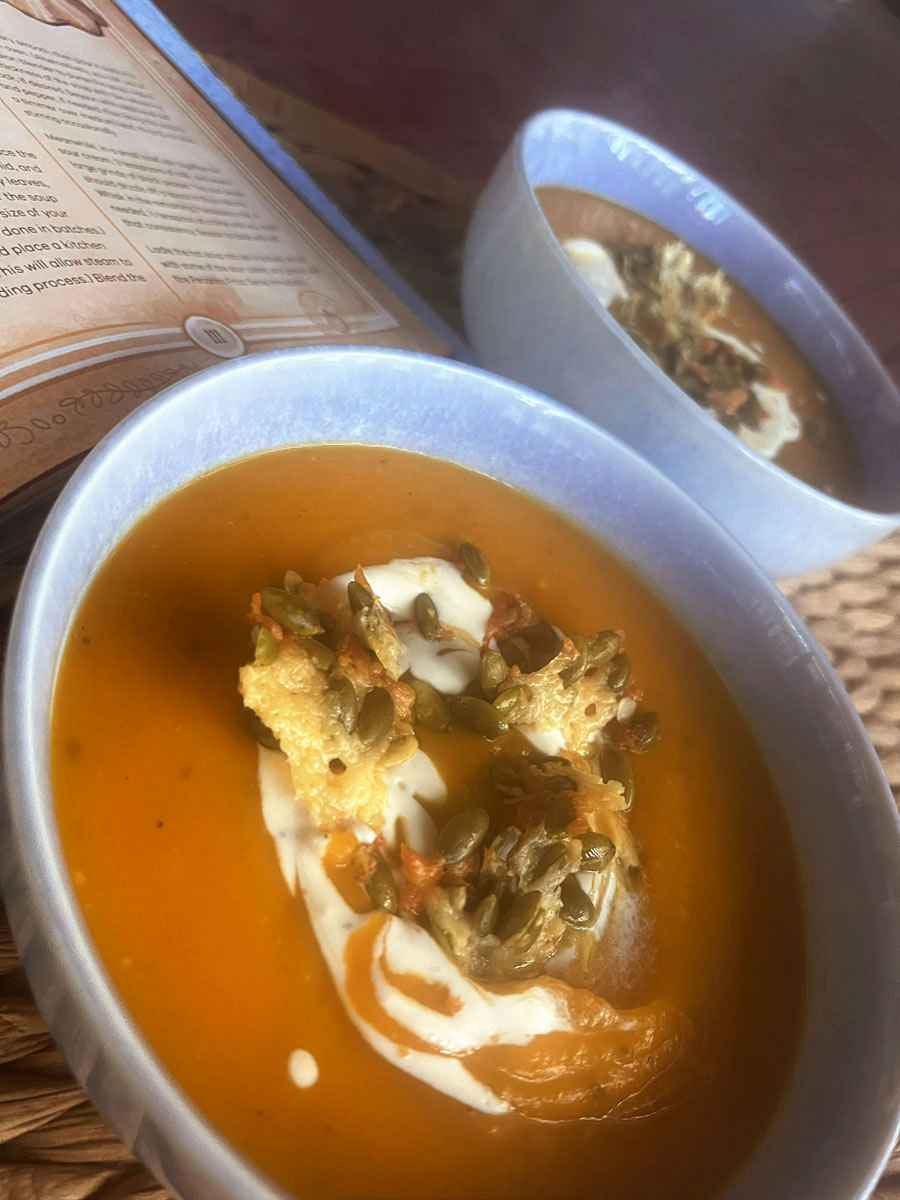 Two bowls of pumpkin soup showing a swirl of cream and pecorino frico in detail, with a corner of the open cookbook visible in the background