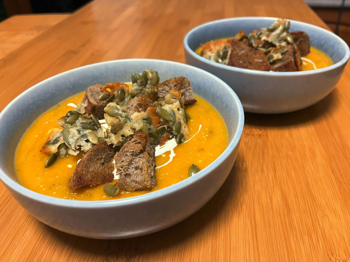 Two blue bowls filled with orange pumpkin soup, with croutons and pieces of crispy fried cheese & pumpkin seeds topping each of them