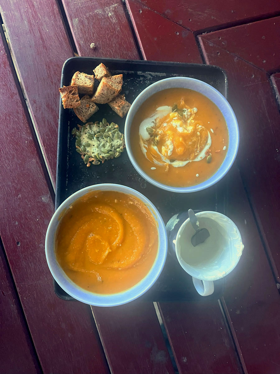 Two blue bowls containing orange pumpkin soup. One has a swirl of cream and flakes of crispy fried cheese in it; the other is plain. Beside them are a cup of cream, a large piece of fried cheese & pumpkin seeds, and some croutons